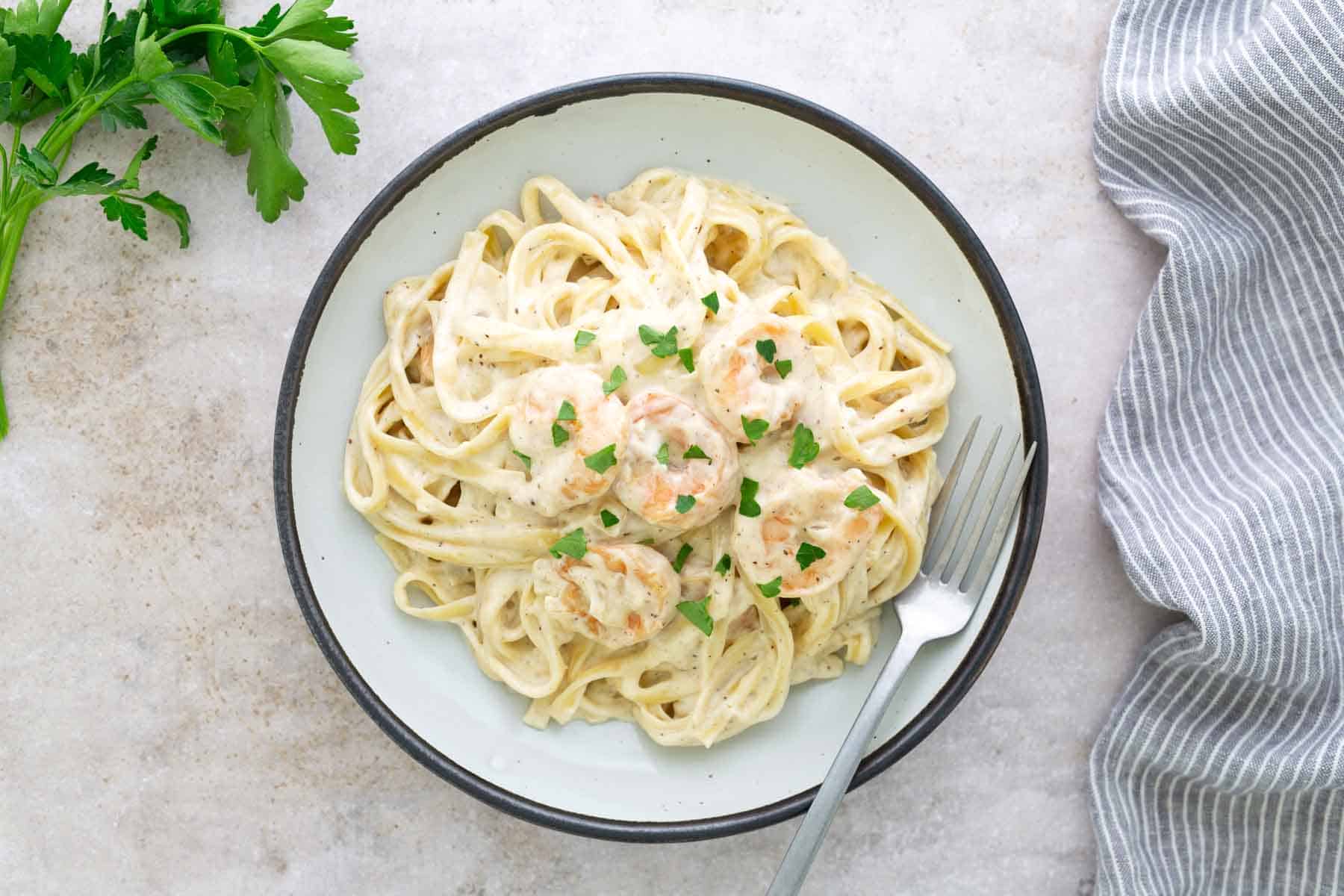 A plate of creamy pasta with shrimp, garnished with parsley, next to a fork and a sprig of fresh parsley on the side.