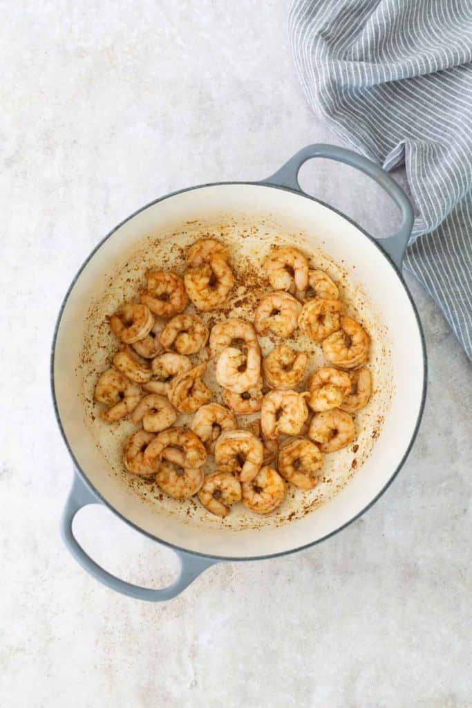 Shrimp sautéed in spices in a light-colored pot on a pale surface, with a blue and white striped cloth beside it.