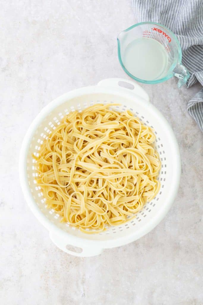 A white colander filled with cooked fettuccine pasta sits on a light surface. A glass measuring cup with reserved pasta water is nearby, next to a blue striped cloth.