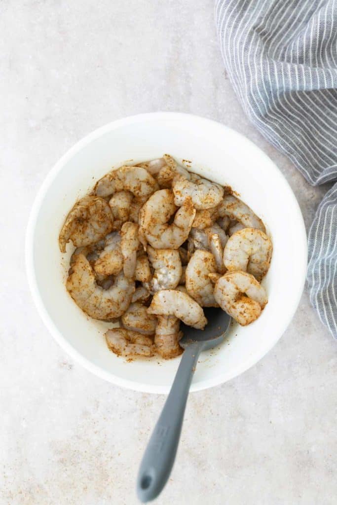 White bowl with raw shrimp seasoned with spices. A gray spoon is placed in the bowl. A striped cloth is visible beside it.