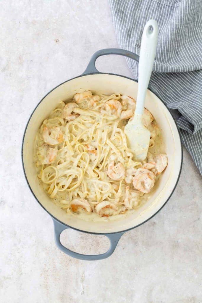 Pot of creamy pasta with sliced sausage and a spatula on a light surface, next to a striped cloth.