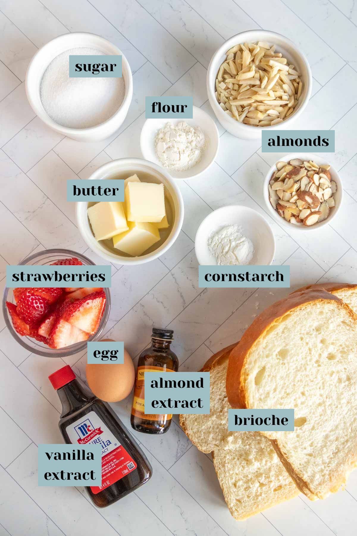 Labeled ingredients on a surface include sugar, flour, almonds, butter, cornstarch, strawberries, egg, almond extract, vanilla extract, and brioche.