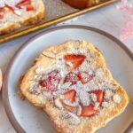 A slice of toast topped with sliced strawberries, almond flakes, and powdered sugar on a plate. More toast slices are visible on a tray in the background.