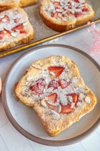 A slice of toast topped with sliced strawberries, almond flakes, and powdered sugar on a plate. More toast slices are visible on a tray in the background.