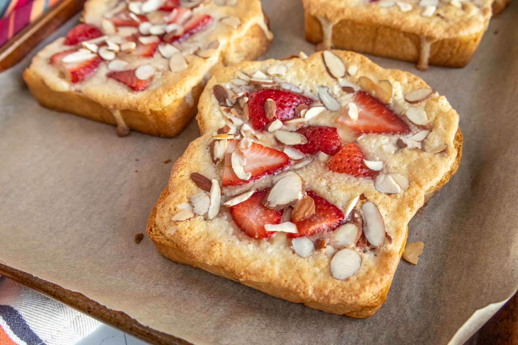 Toasted bread topped with sliced strawberries, almond slivers, and a light glaze on a parchment-lined baking sheet.