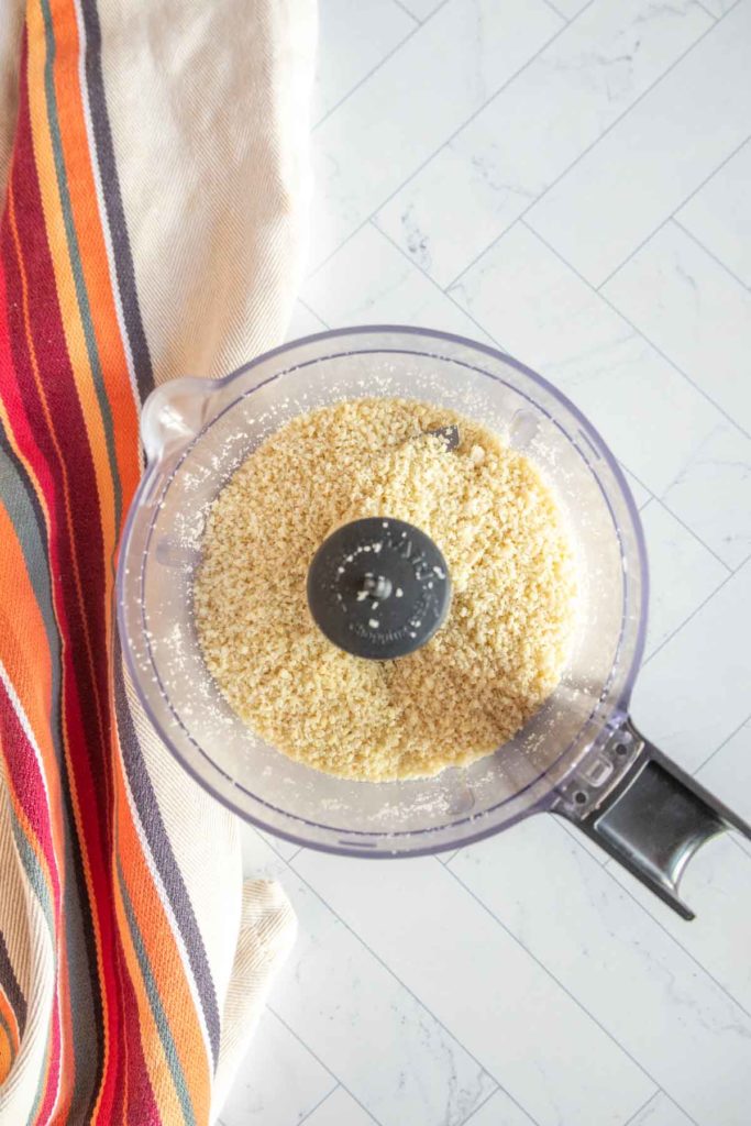 A food processor containing ground nuts next to a striped cloth on a tiled surface.