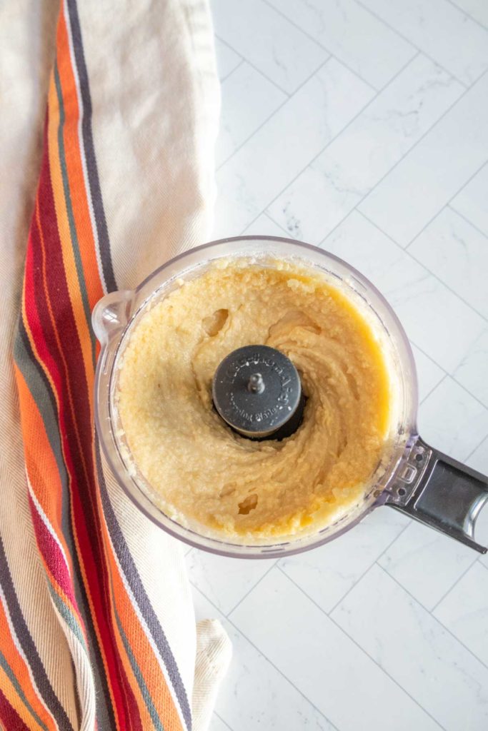 Food processor with blended beige mixture next to a colorful striped cloth on a tiled surface.