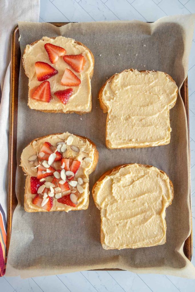 Four slices of bread with cream spread on a tray; two topped with sliced strawberries, one with strawberries and almonds, and one plain.