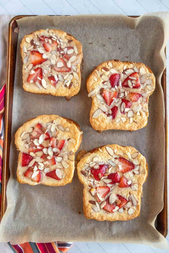 Four slices of toast topped with sliced strawberries and almond slivers on a parchment-lined baking tray.