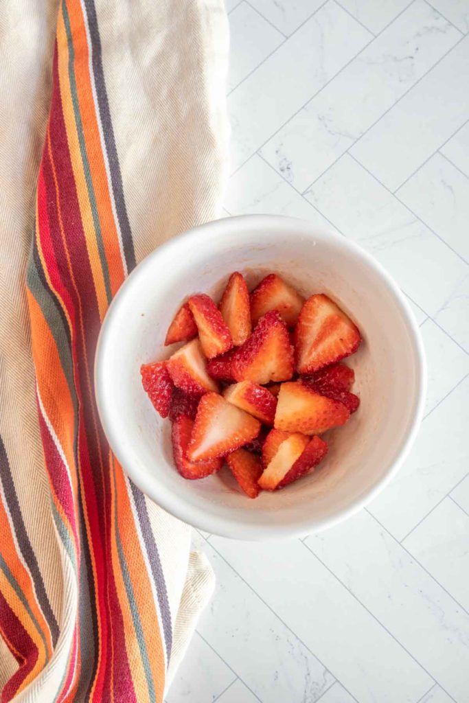 White bowl with sliced strawberries next to a striped cloth on a tiled surface.