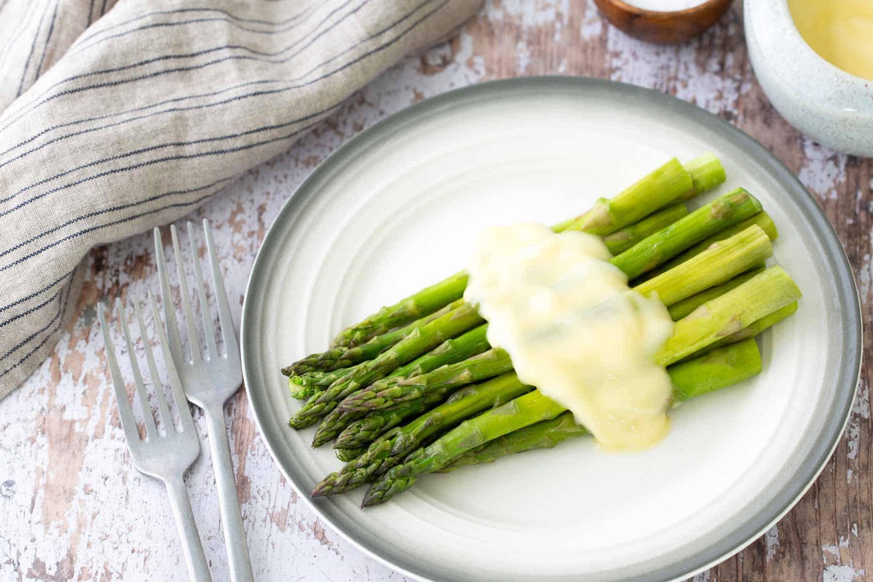 A plate of steamed asparagus topped with hollandaise sauce. Nearby are two forks and a striped napkin on a rustic wooden surface.