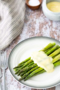 A plate of steamed asparagus topped with a creamy sauce, placed on a rustic wooden table next to silverware and a striped cloth.