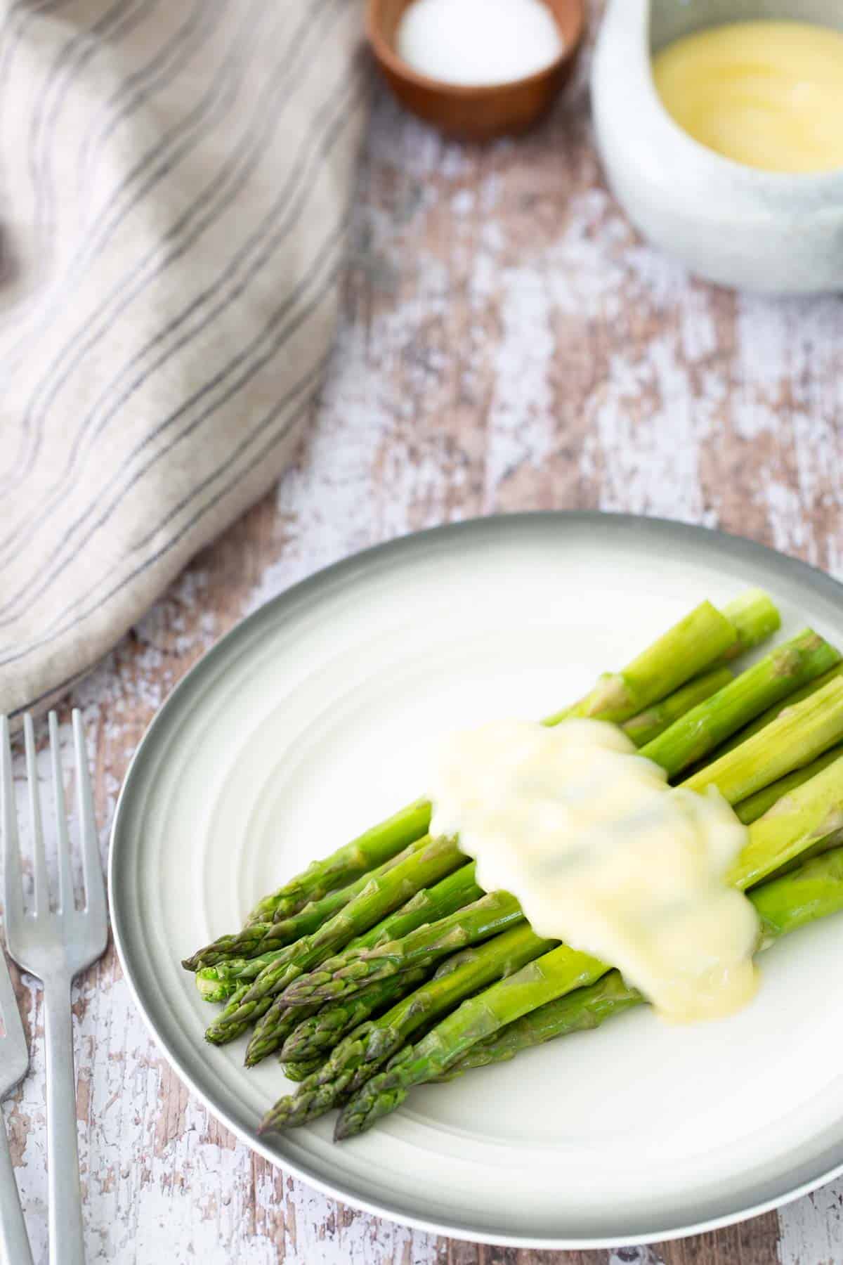 A plate of steamed asparagus topped with a creamy sauce, placed on a rustic wooden table next to silverware and a striped cloth.