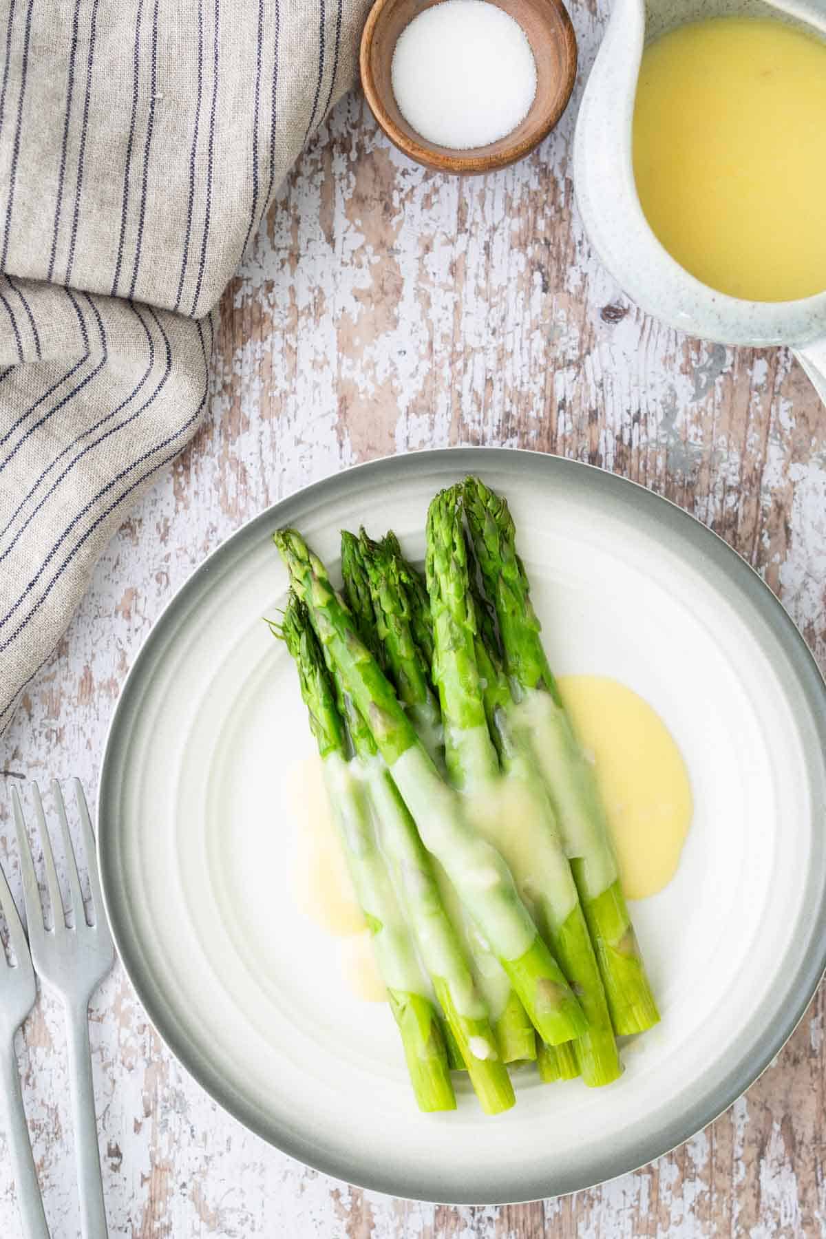 Plate of steamed asparagus spears drizzled with hollandaise sauce on a rustic table setting with salt, fork, knife, napkin, and a jug of sauce.