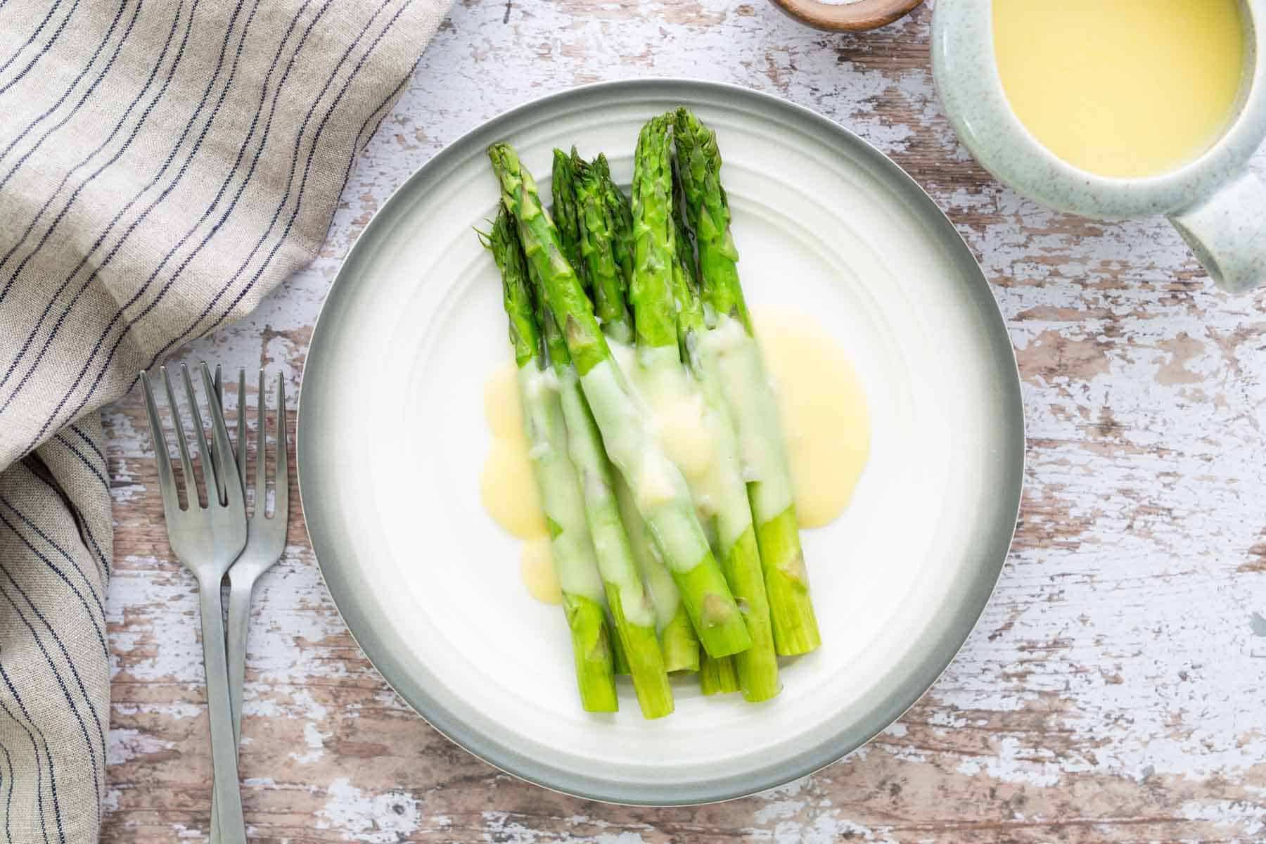 Plate of steamed asparagus topped with a creamy sauce, placed on a white plate with a fork and a striped cloth nearby.