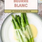 A plate of asparagus topped with beurre blanc sauce, with a cloth napkin and a bowl of yellow sauce in the background.