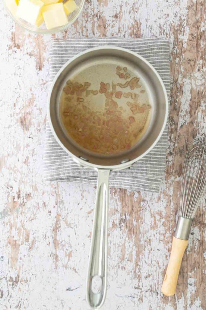 Sliced onions sautéing in a saucepan on a striped cloth, with a bowl of butter cubes and a whisk nearby on a rustic wooden surface.