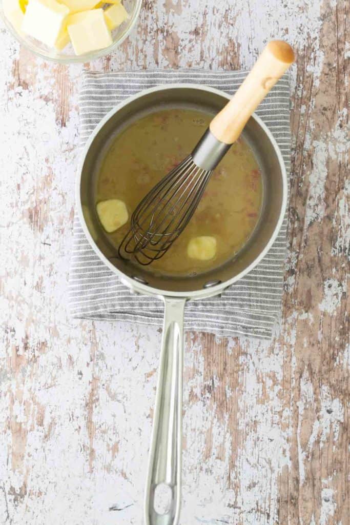 A saucepan with a whisk and melting butter cubes sits on a striped cloth. Nearby is a bowl with more butter cubes on a rustic wooden surface.