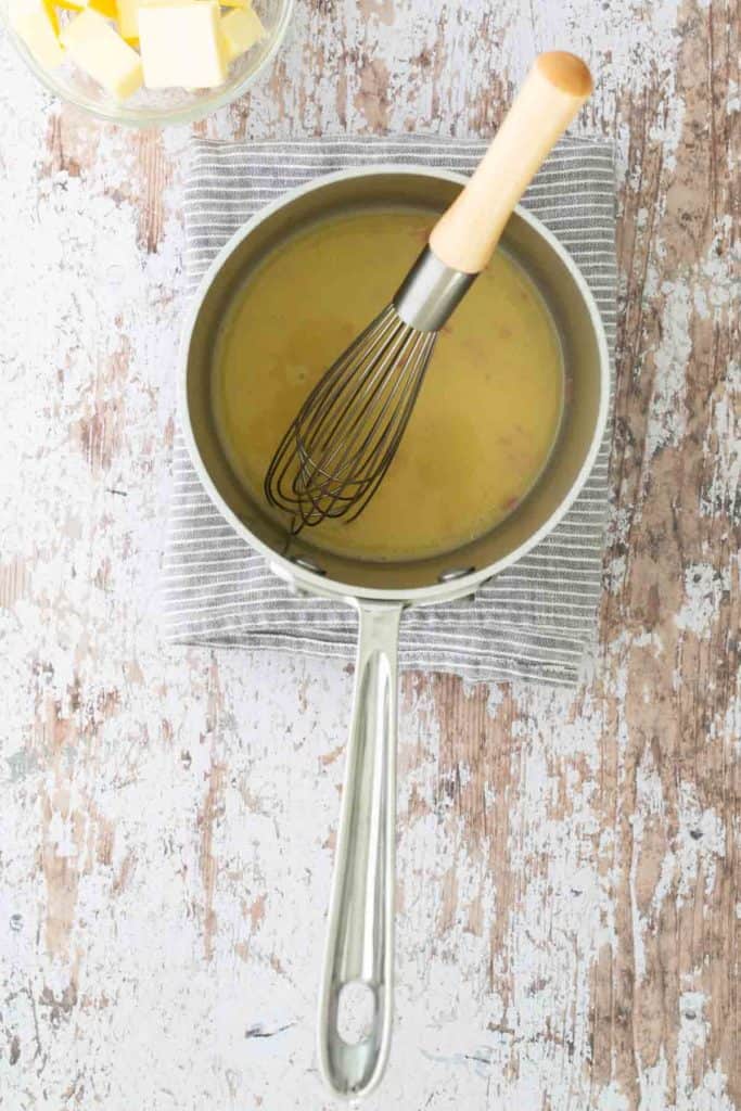 A whisk in a saucepan filled with yellow sauce, placed on a striped cloth on a rustic wooden surface. A bowl with butter cubes is visible in the corner.