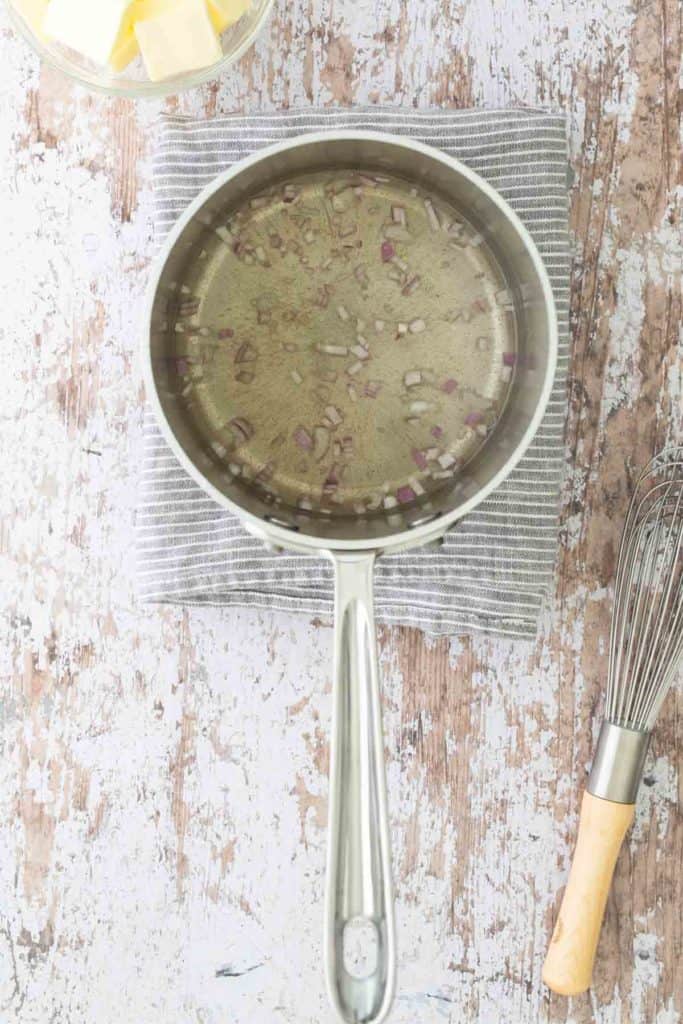 A saucepan with chopped onions in liquid on a striped cloth, next to a block of butter in a dish and a whisk on a rustic wooden surface.