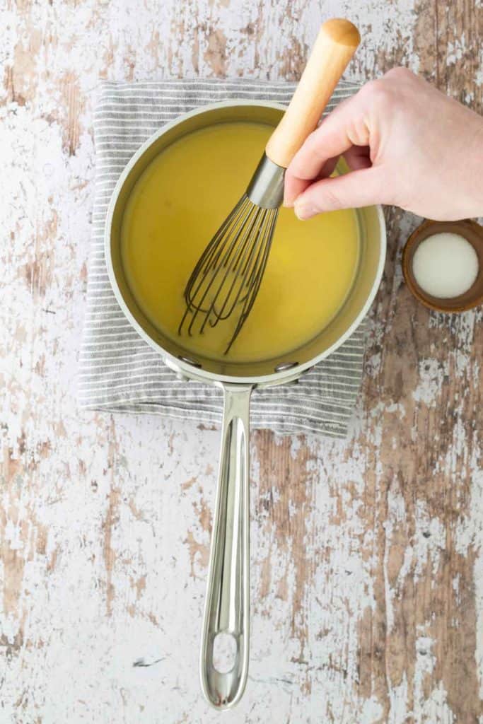 A person's hand uses a whisk in a saucepan with yellow liquid on a striped cloth, next to a small bowl of salt on a wooden surface.