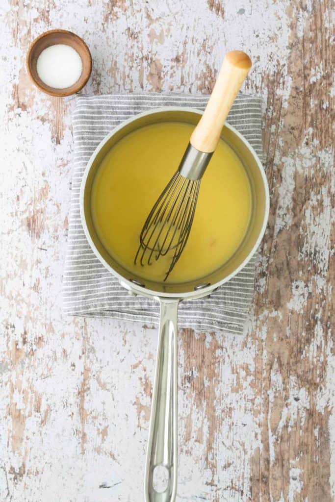 A saucepan filled with yellow liquid and a whisk sits on a striped cloth, on a rustic wooden surface. A small bowl of salt is nearby.