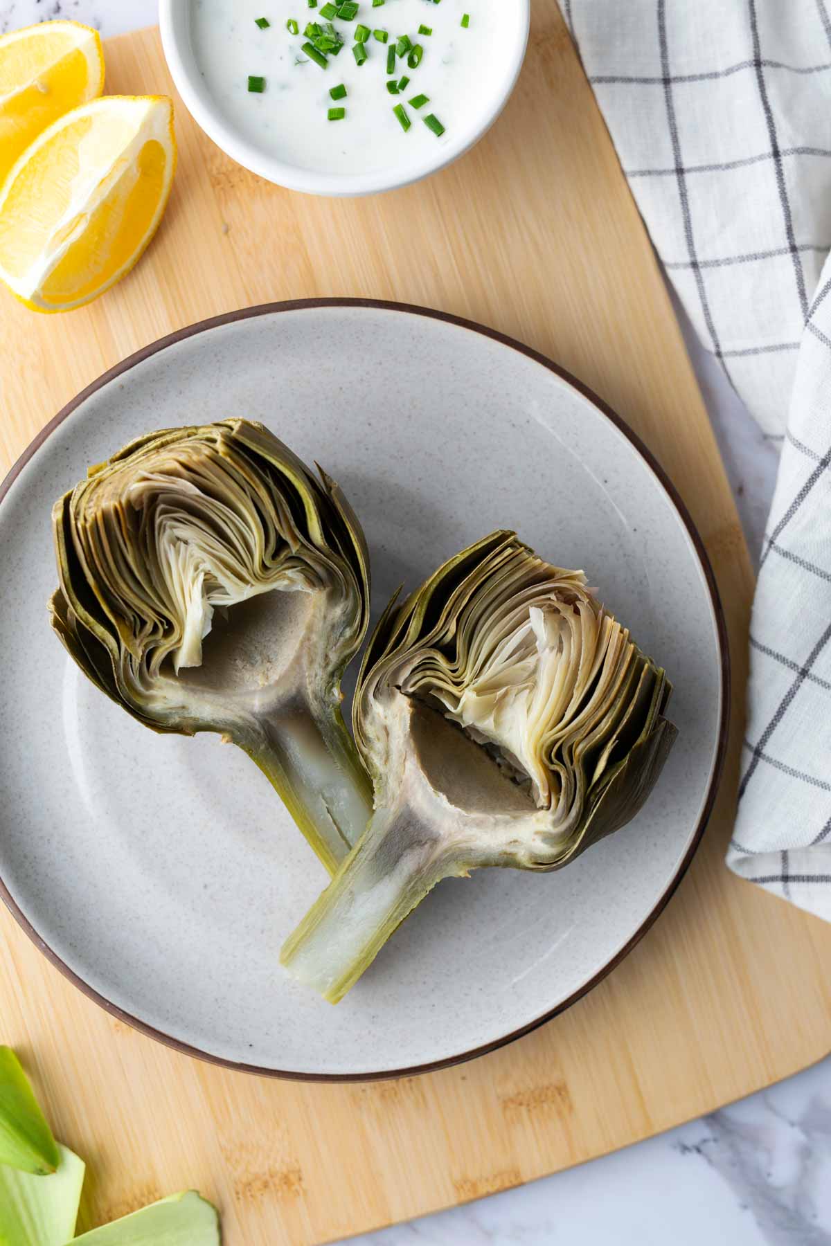 Two halved artichokes on a plate with a small bowl of dipping sauce, lemon wedge, and cloth napkin on a wooden board.