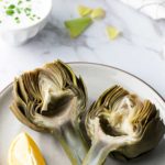 A plate with two artichoke halves and a lemon wedge. A bowl of dipping sauce with chopped green herbs is in the background.