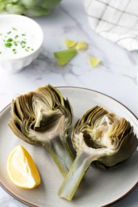 A plate with two artichoke halves and a lemon wedge. A bowl of dipping sauce with chopped green herbs is in the background.