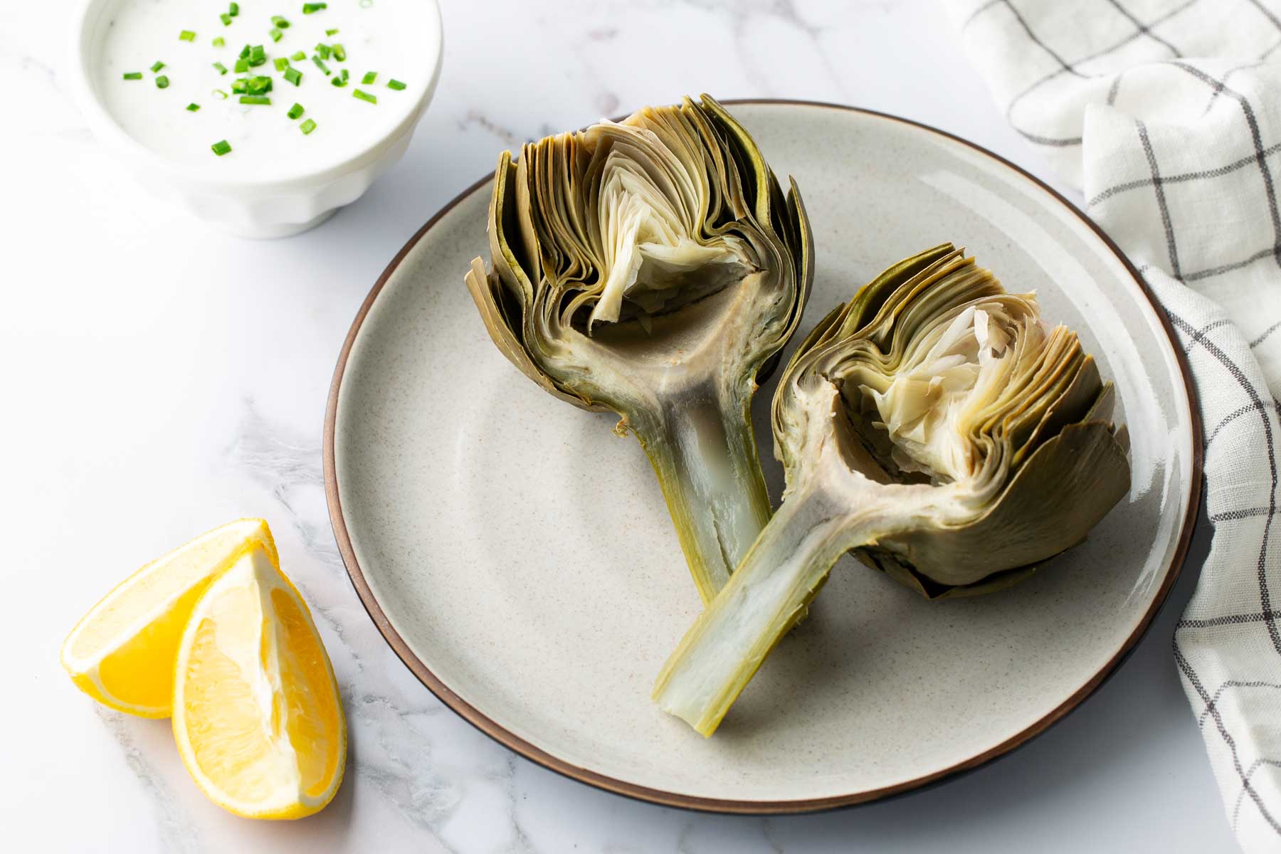 Two halved artichokes on a plate with lemon wedges and a small bowl of dipping sauce, garnished with chives.