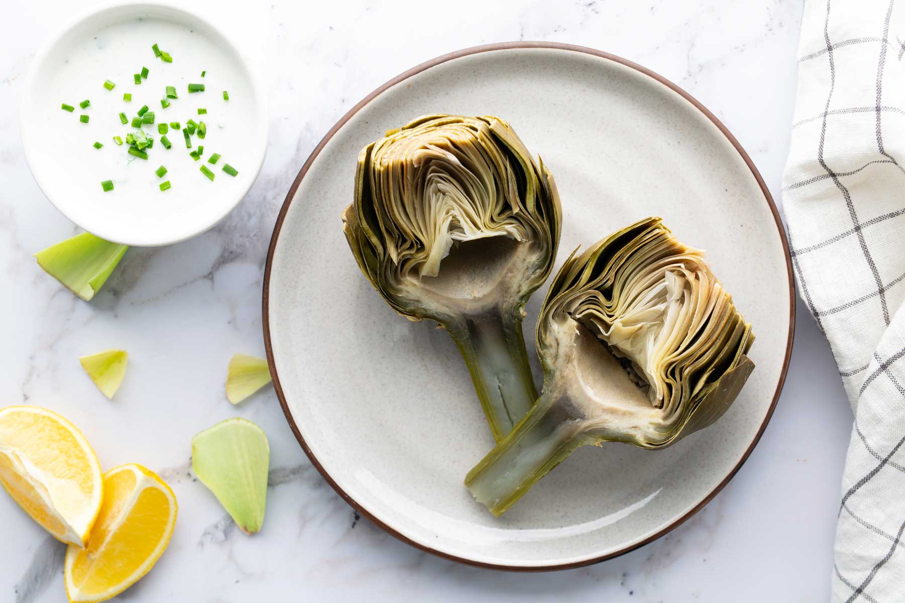 Two halved artichokes on a plate with a small bowl of creamy dip and lemon wedges on a marble surface. A checkered cloth is beside the plate.