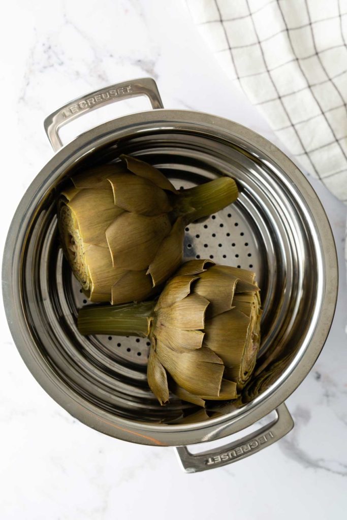 Two artichokes in a stainless steel steamer pot on a marble countertop, with part of a checkered cloth visible.