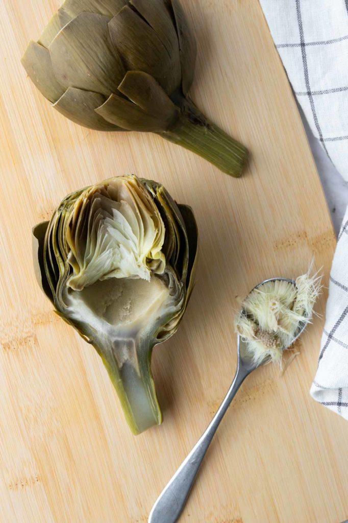 Artichoke on a wooden board, one whole and one halved showing the interior. A spoon with removed choke is nearby. A striped cloth is partially visible on the right.