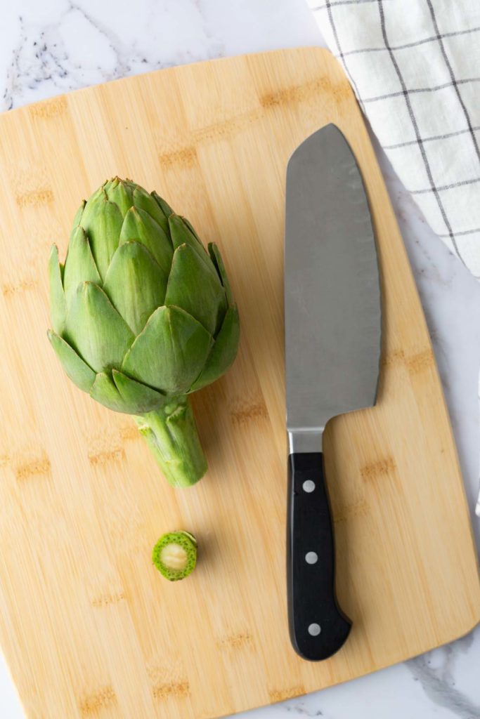 An artichoke and a chef's knife on a wooden cutting board, with a small slice of the artichoke stem nearby.