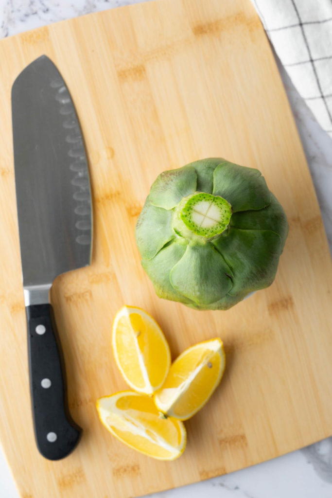 Artichoke and lemon wedges on a wooden cutting board with a chef's knife.