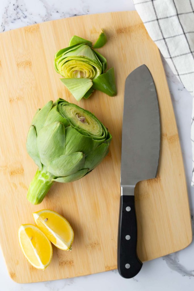 Artichoke and lemon wedges on a wooden cutting board with a knife; artichoke has been partially sliced.