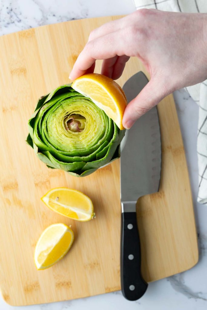 A hand holds a lemon wedge over a halved artichoke on a wooden cutting board. A knife and additional lemon wedges are nearby.