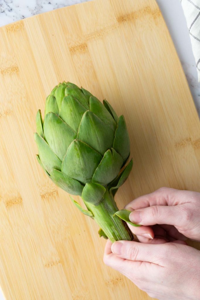 A person peeling the leaves of a fresh artichoke on a wooden cutting board.