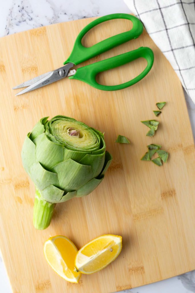 Artichoke on a wooden cutting board with trimmed leaves, lemon wedges, and green-handled scissors nearby.