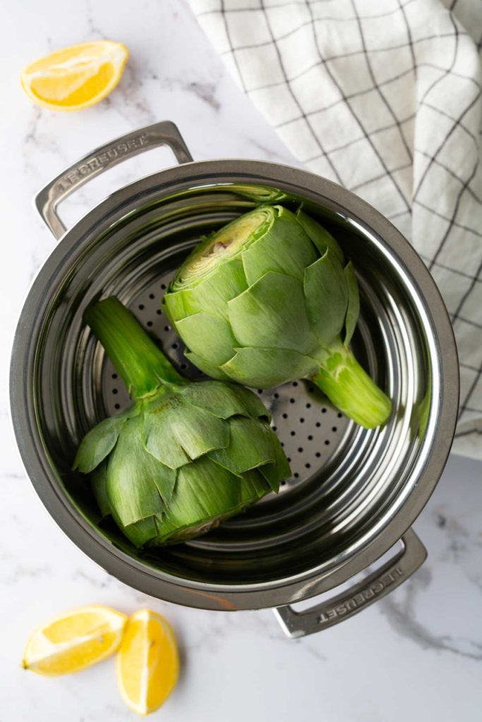 Two artichokes in a stainless steel steamer pot, placed on a marble surface. Lemon wedges are nearby, and a checkered cloth is partially visible.