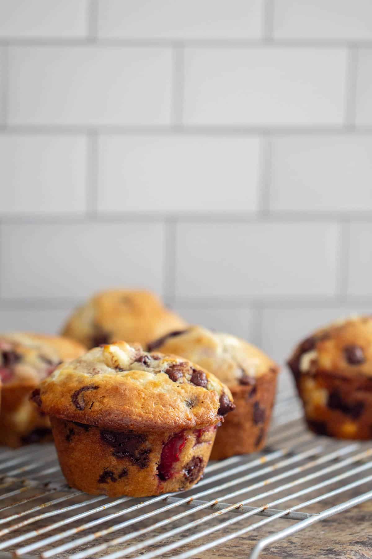 Chocolate chip muffins with strawberries on a cooling rack in front of a white tile wall.