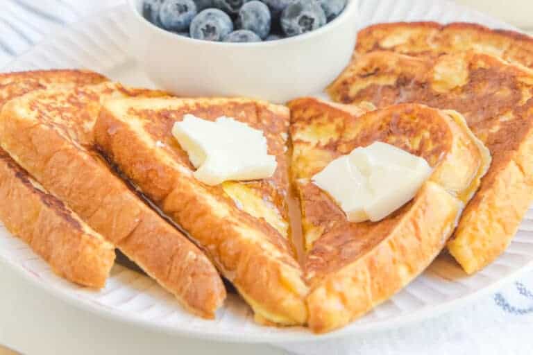 Plate of golden brown French toast slices with butter on top, served with a bowl of fresh blueberries.