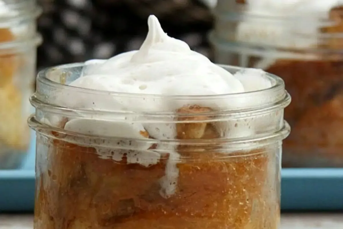 A glass jar filled with bread pudding, topped with a swirl of whipped cream, sits on a blue tray with similar jars in the background.